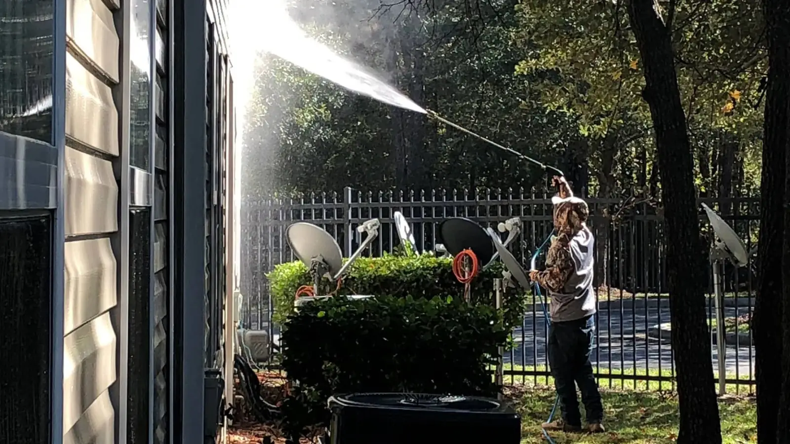 Pressure washing service in progress on an exterior building facade and surrounding areas, with a technician actively spraying surfaces near a fenced property.
